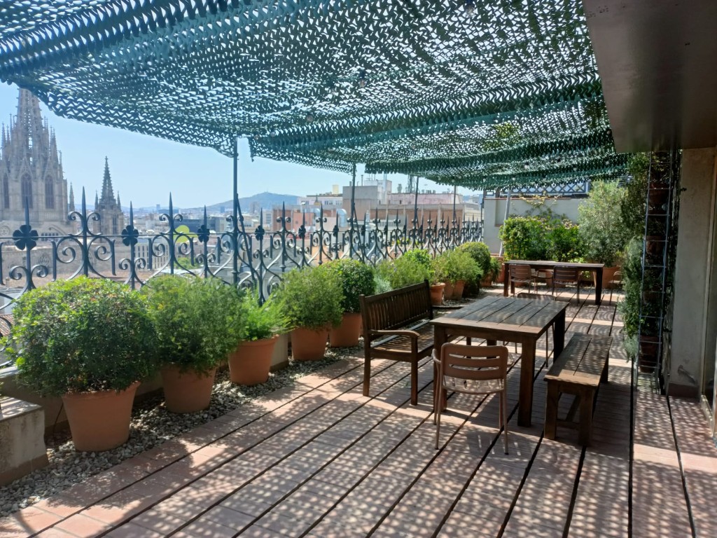 Wide view of Pier07 terrace with outdoor table, bench, shade net, and Barcelona skyline